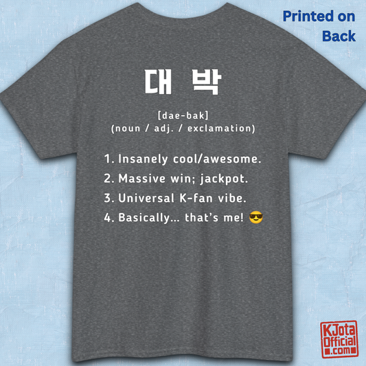 A gray short-sleeve T-shirt flatlay shown from the back on a light blue textured background, with white printed Korean text and a dictionary-style definition of Daebak 대박 Korean slang. K-fandom merch by K-Jota
