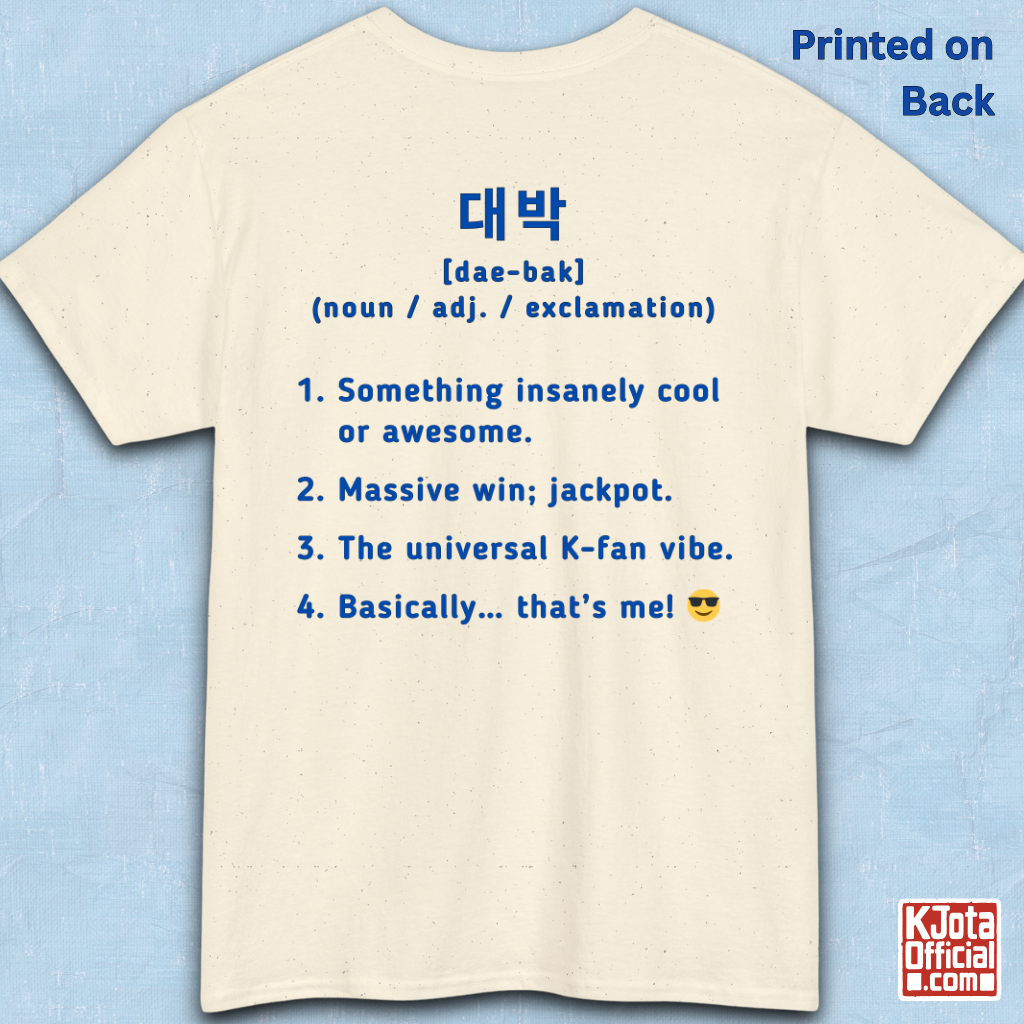 A beige short-sleeve T-shirt flatlay shown from the back on a light blue textured background, with blue printed Korean text and a dictionary-style definition of Daebak 대박 Korean slang. K-fandom merch by K-Jota