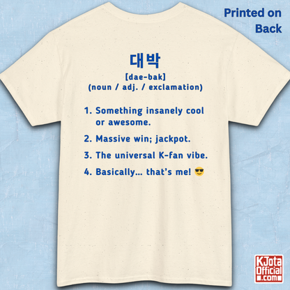 A beige short-sleeve T-shirt flatlay shown from the back on a light blue textured background, with blue printed Korean text and a dictionary-style definition of Daebak 대박 Korean slang. K-fandom merch by K-Jota