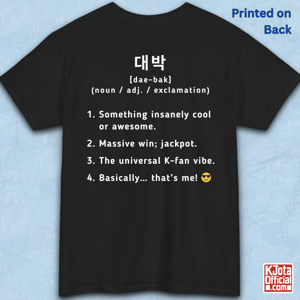 A black short-sleeve T-shirt flatlay shown from the back on a light blue textured background, with white printed Korean text and a dictionary-style definition of Daebak 대박 Korean slang. K-fandom merch by K-Jota