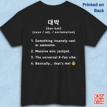 A black short-sleeve T-shirt flatlay shown from the back on a light blue textured background, with white printed Korean text and a dictionary-style definition of Daebak 대박 Korean slang. K-fandom merch by K-Jota