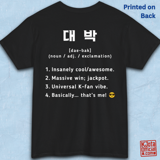 A black short-sleeve T-shirt flatlay shown from the back on a light blue textured background, with white printed Korean text and a dictionary-style definition of Daebak 대박 Korean slang. K-fandom merch by K-Jota
