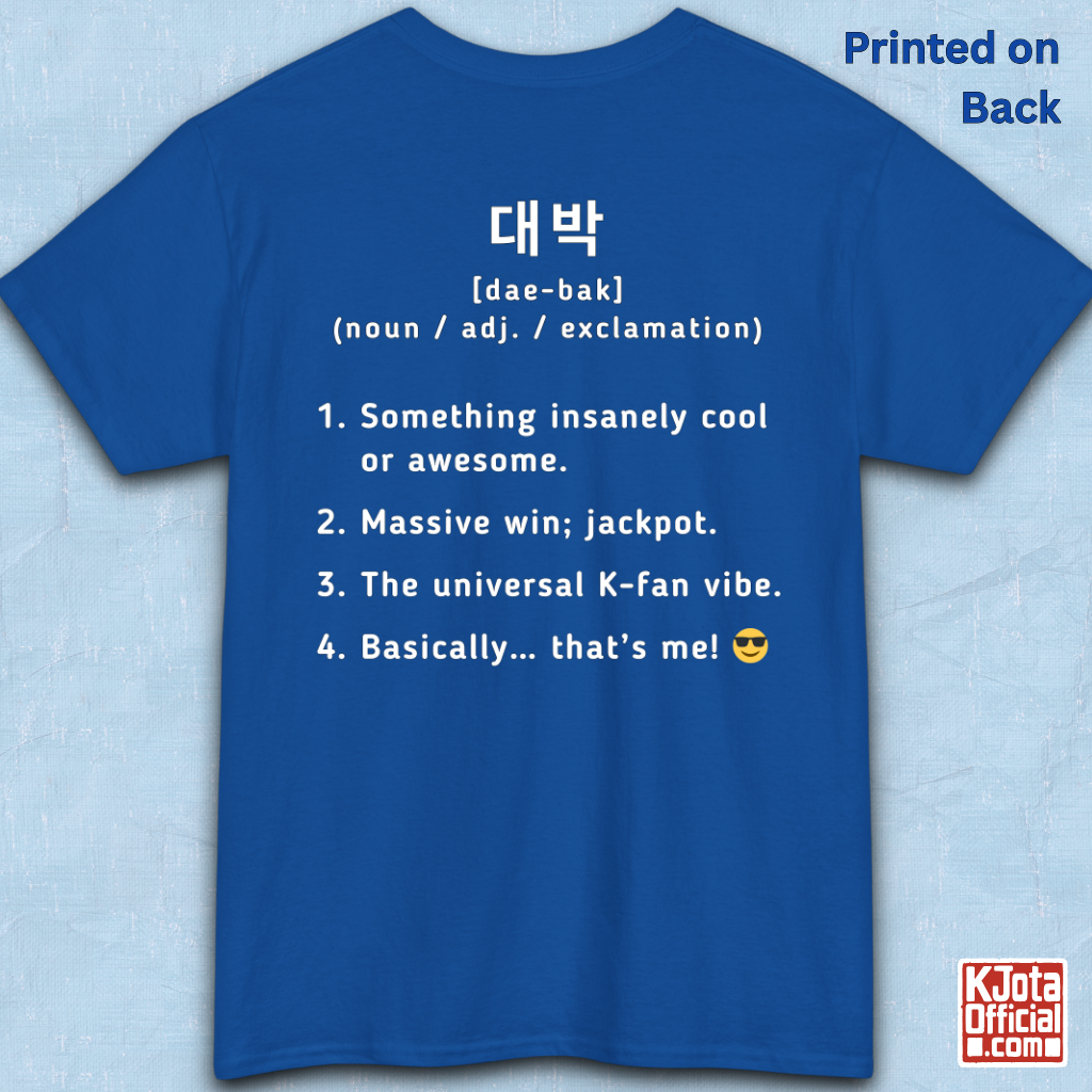 A blue short-sleeve T-shirt flatlay shown from the back on a light blue textured background, with white printed Korean text and a dictionary-style definition of Daebak 대박 Korean slang. K-fandom merch by K-Jota