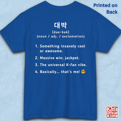 A blue short-sleeve T-shirt flatlay shown from the back on a light blue textured background, with white printed Korean text and a dictionary-style definition of Daebak 대박 Korean slang. K-fandom merch by K-Jota