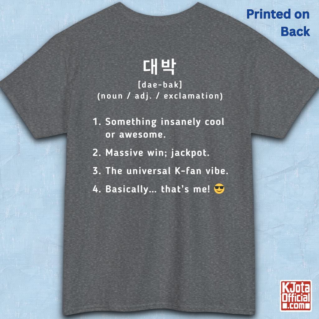 A gray short-sleeve T-shirt flatlay shown from the back on a light blue textured background, with white printed Korean text and a dictionary-style definition of Daebak 대박 Korean slang. K-fandom merch by K-Jota