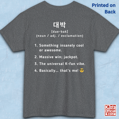 A gray short-sleeve T-shirt flatlay shown from the back on a light blue textured background, with white printed Korean text and a dictionary-style definition of Daebak 대박 Korean slang. K-fandom merch by K-Jota