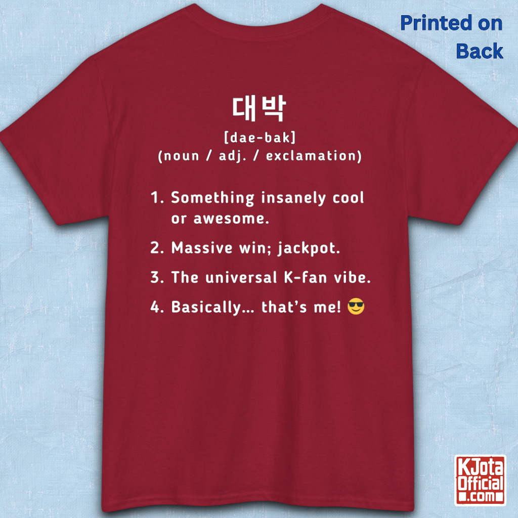 A red short-sleeve T-shirt flatlay shown from the back on a light blue textured background, with white printed Korean text and a dictionary-style definition of Daebak 대박 Korean slang. K-fandom merch by K-Jota