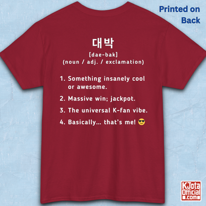 A red short-sleeve T-shirt flatlay shown from the back on a light blue textured background, with white printed Korean text and a dictionary-style definition of Daebak 대박 Korean slang. K-fandom merch by K-Jota