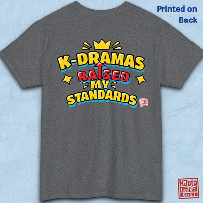 'K-DRAMAS RAISED MY STANDARDS' yellow, red, and blue lettering with an uparrow in place of letter “I” printed on a gray t-shirt backside flatlay. Realistic expectations K-fan graphic t-shirt by K-Jota