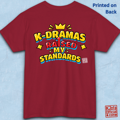 'K-DRAMAS RAISED MY STANDARDS' yellow, red, and blue lettering with an uparrow in place of letter “I” printed on a red t-shirt backside flatlay. Realistic expectations K-fan graphic t-shirt by K-Jota