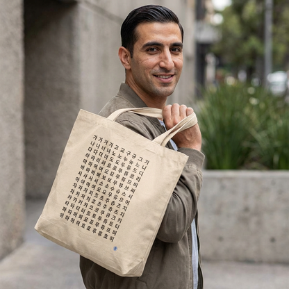 man holding Hangeul alphabet chart featuring Korean consonants and vowels on beige canvas tote bag by Kjota