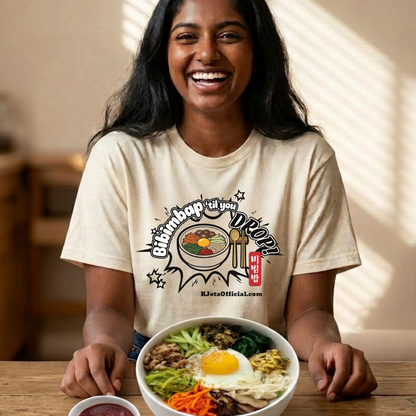 A woman wearing a beige t-shirt by K-Jota showing Korean food mixed rice bowl and sitting at a table with a bowl of bibimbap. The graphic features a hand-illustrated bowl of bibimbap with a spoon and chopsticks, and texts “Bibimbap ‘til You Drop 비빔밥” and “KJotaOfficial.com”.