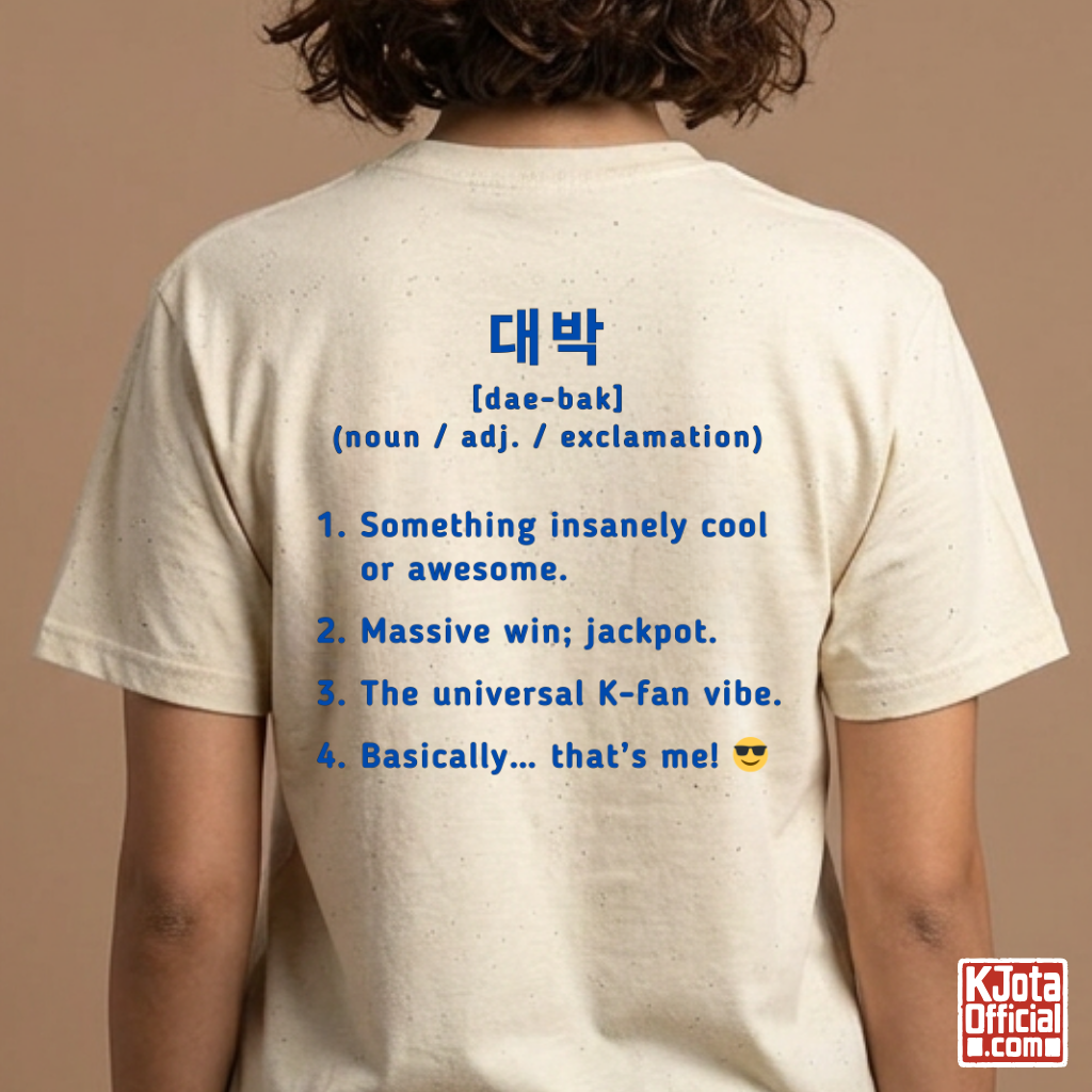 woman wearing a beige short-sleeve T-shirt shown from the back with blue printed Korean text and a dictionary-style definition of Daebak 대박 Korean slang. K-fandom merch by K-Jota
