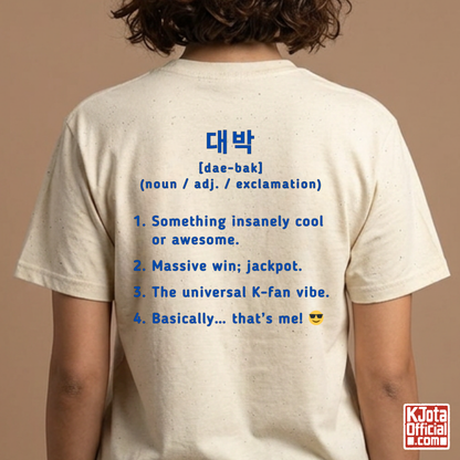 woman wearing a beige short-sleeve T-shirt shown from the back with blue printed Korean text and a dictionary-style definition of Daebak 대박 Korean slang. K-fandom merch by K-Jota