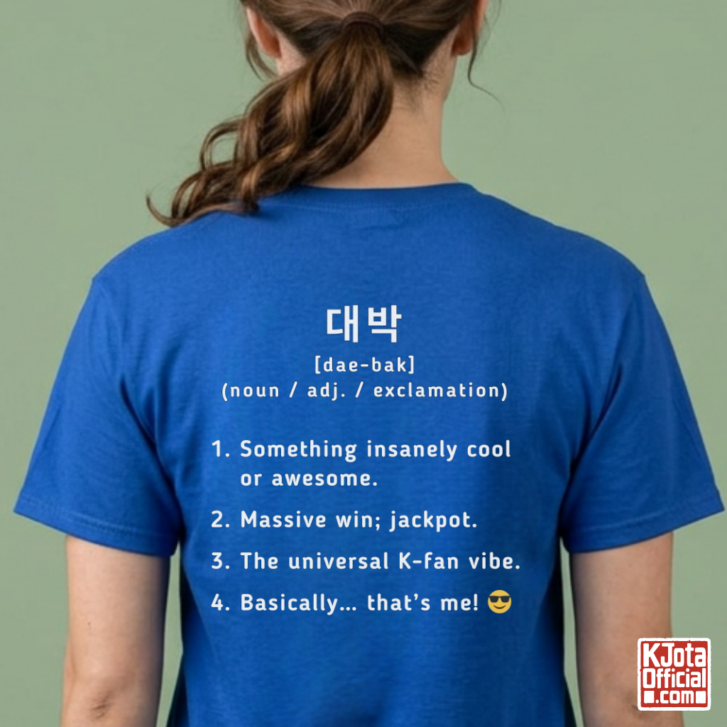 woman wearing a gray short-sleeve T-shirt shown from the back with white printed Korean text and a dictionary-style definition of Daebak 대박 Korean slang. K-fandom merch by K-Jota