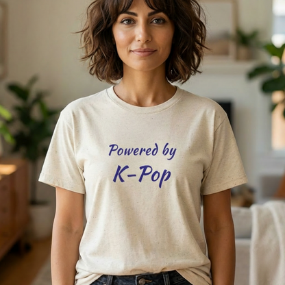 A woman wearing a light beige crewneck t-shirt with the text 'Powered by K-Pop' printed in blue on the front, set against a softly blurred indoor background by Kjota